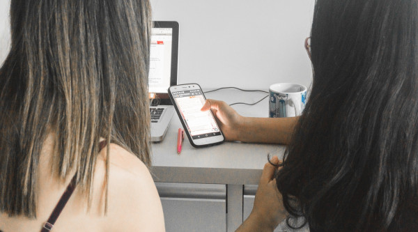 Zwei Frauen sitzen vor einem Computer und schauen auf ein Mobilphone