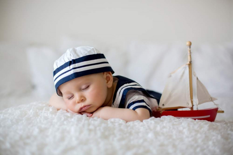 Ein Baby in Matrosen-Outfit schläft auf einer Decke, neben ihm ein Spielzeug-Segelboot. 