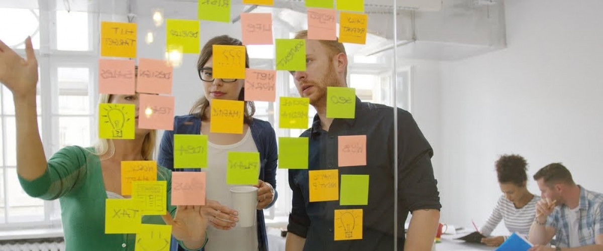 An einer Glaswand sind Post-its befestigt. Davor stehen 3 junge Menschen und diskutieren die Ideen. 